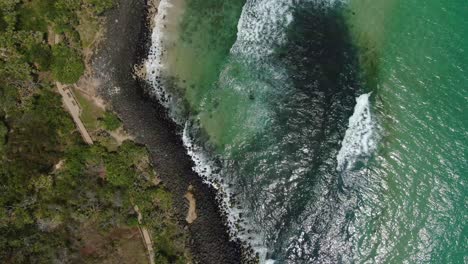 Looking-vertically-at-small-waves-breaking-on-rocky-beach-slight-spin,-bright-summers-day,-drone