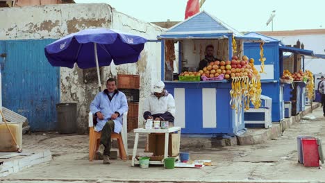 Escena-De-4k-Uhd-De-Dos-Viejos-Pescadores-Sentados-Bajo-Un-Paraguas-En-La-Zona-Del-Puerto-De-La-Antigua-Ciudad-Marroquí-De-Essaouira-Con-Un-Puesto-De-Frutas-En-El-Fondo-Y-Un-Gato-Caminando