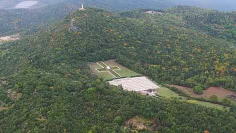 Flight-above-cross-at-famous-Polish-cemetery-at-Monte-Cassino-with-green-trees-on-hillside-forest,-Italy,-overhead-aerial-approach
