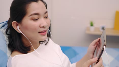 Cheerful-young-beautiful-Asian-woman-relaxing-and-listening-to-music-by-tablet-with-headphones-on-the-bed-at-home-lifestyle-and-pleasure-concept