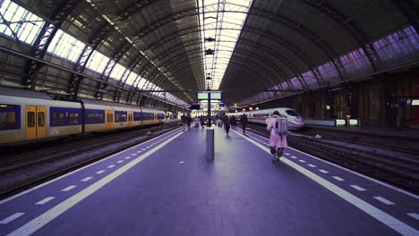 Viajeros-Caminando-A-Lo-Largo-De-La-Plataforma-Con-Tren-Saliendo-En-La-Estación-De-Amsterdam,-Cámara-Lenta
