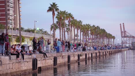 Personas-Sentadas-Cerca-Del-Agua-En-El-Puerto-Con-Máscaras-Faciales---Noche-En-Málaga,-España