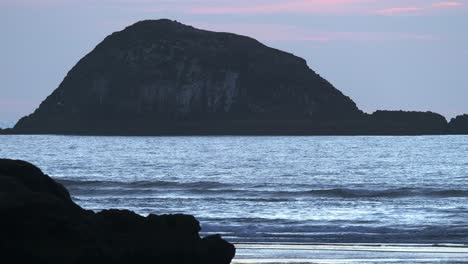 Wilde-Seevögel,-Die-über-Ruhigen-Ozean-Fliegen,-Mit-Plätscherndem-Wasser-Und-Bergsilhouette-Im-Hintergrund-Am-Muriwai-Beach,-Auckland,-Neuseeland