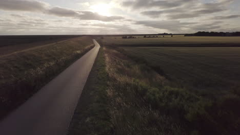 fast-drone-flight-forward-above-an-empty-dyke-road,-back-light,-scenic-landscape,-sunset,-sky-with-scenic-clouds