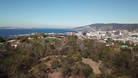 Aerial-pan-up-over-trees-to-reveal-seaside-port-and-city-of-Hobart,-Tasmania,-Australia