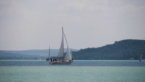 Velero-En-El-Lago-Balaton-Con-Tihany-Y-El-Viaducto-De-Kőröshegy-Al-Fondo,-Filmado-En-Cámara-Lenta-De-180-Fps-En-Balatonfüred