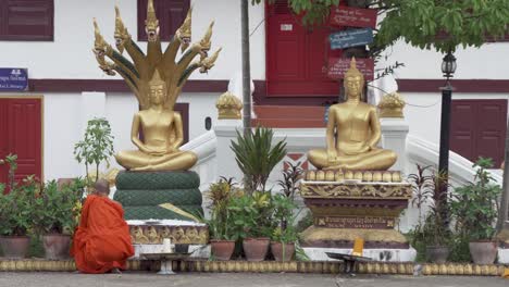 Monje-Con-Túnica-Naranja-Reza-Dentro-Del-Templo-Budista-Con-Dos-Estatuas-Doradas-De-Buda-En-Laos,-Luang-Prapang