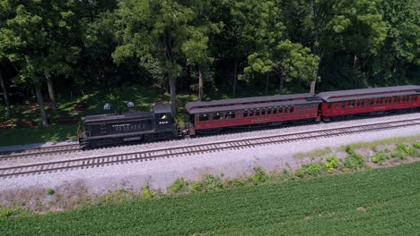 Una-Vista-Aérea-De-Una-Locomotora-Diésel-Tirando-De-Coches-De-Pasajeros-Antiguos-A-Través-De-La-Campiña-Amish