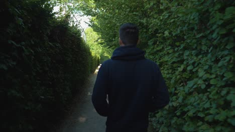 Man-wearing-blue-hoodie-walking-in-lush-green-park,-back-tracking-shot