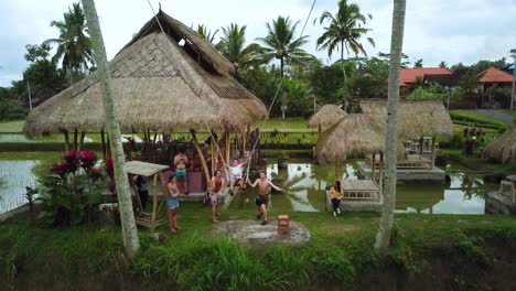 Drone-Shot-of-a-tourist-enjoying-a-swing-between-two-coconut-trees-that-swings-out-over-some-Rice-Terraces-in-Bali,-Indonesia