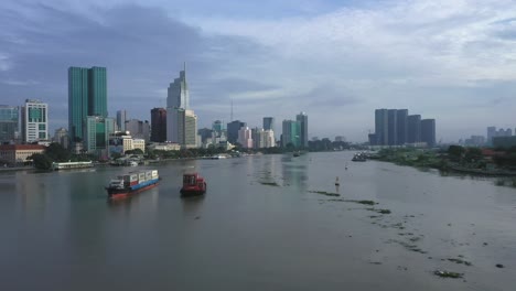 Drohnenaufnahme-Eines-Frachtschiffs-Und-Eines-Kleineren-Schiffs,-Das-Schiffscontainer-Entlang-Des-Saigon-flusses-Transportiert,-Mit-Dem-Finanz--Und-Stadtzentrum-In-Voller-Sichtweite