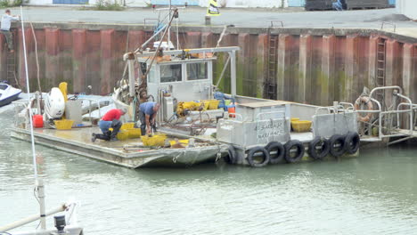 Two-Oyster-producers-who-wash-with-a-water-jet-oysters-and-seashells-on-the-bridge-of-a-boat-after-a-outing-to-sea,-port-of-Saint-Trojan-les-Bain-in-Oléron