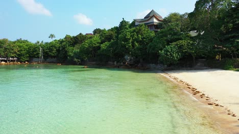 Paradise-holiday-resort-hidden-inside-trees-forest-near-white-sandy-beach-washed-by-calm-clear-water-of-turquoise-lagoon-in-Thailand