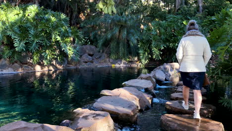 Middle-aged-women-walks-along-stone-pathway-over-flowing-water-from-waterfall,-Toowoomba-Queensland