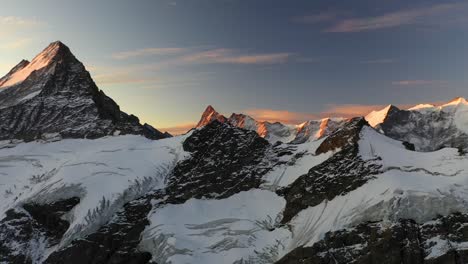Schöne-Und-Langsame-Drohnenaufnahme-Der-Verschneiten-Landschaft,-Die-Die-Alpen-Der-Schweiz-Während-Des-Sommers-Im-Farbenfrohen-Und-Dramatischen-Sonnenaufgang-Bedeckt