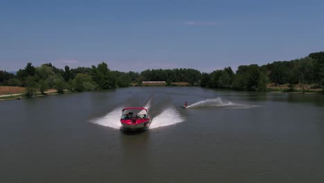 Male-Slalom-Water-Skier-Towed-by-Red-Ski-Boat-at-Ski-Park-Lake-AERIAL