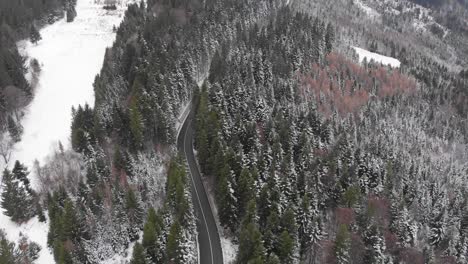 Aerial,-tilt-up,-drone-shot,-of-a-road-in-middle-of-snowy-spruce-forest,-on-a-mountain,-on-a-cloudy,-winter-day,-in-Romania