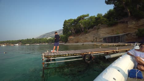 Young-man-standing-on-end-of-wooden-dock-as-small-white-boat-with-woman-in-dress-arrives