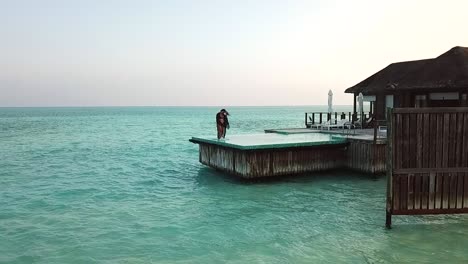 Aerial,-slow-motion,-drone-shot,-of-a-young-woman,-walking-on-the-glass-covered-pool,-at-a-bungalow-on-the-sea,,-on-a-sunny-day,-on-the-Conrad-Rangali-island,-in-Maldives