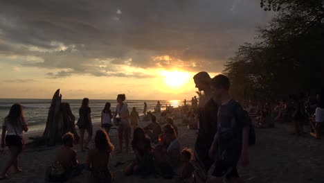 Large-group-silhouette-people-gathered-on-sunset-glowing-sandy-beach-shoreline-watching-ocean