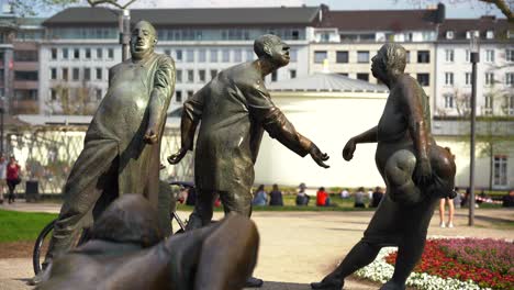 Estatua-De-Bronce-Con-Gente-Y-Piscina-De-Hidromasaje-En-Aquisgrán,-Alemania,-Llamada-Circulación-De-Dinero-Con-El-Elisenbrunnen-En-Segundo-Plano