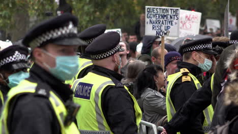 Eine-Reihe-Von-Polizisten-Mit-Schützenden-Gesichtsmasken-Stehen-Und-Beobachten-Einen-Protest-Gegen-Das-Coronavirus-Und-Die-Qanon-Verschwörung,-Während-Ein-Demonstrant-Ein-Plakat-Mit-Der-Aufschrift-„Stellen-Sie-Das-Narrativ-Der-Regierung-In-Frage,-Erheben-Sie-Sich-Jetzt“-In-Der-Hand.