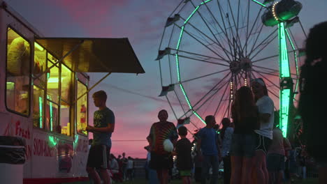 Joven-Comprando-Comida-Del-Puesto-En-Un-Pequeño-Carnaval-Al-Atardecer,-Plano-General