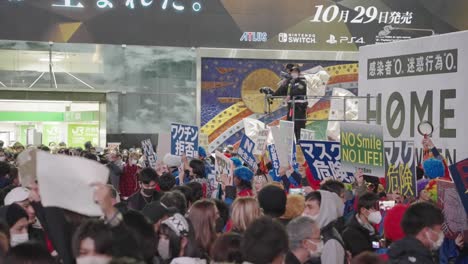 Grupo-De-Personas-Sosteniendo-Pancartas-Protestando-Contra-El-Uso-De-Máscaras-Y-Policías-En-Una-Torre-De-Vigilancia-Observando-En-El-Fondo-Durante-La-Noche-De-Halloween-En-Shibuya,-Tokio,-Japón---Cámara-Lenta