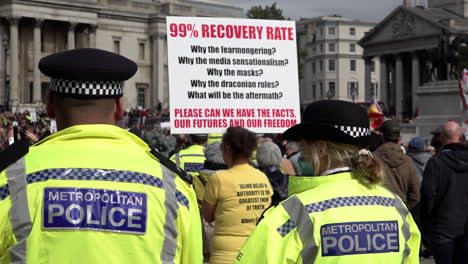 A-slow-motion-shot-of-two-police-officers-wearing-face-masks-monitoring-a-Coronavirus-conspiracy-protest-where-one-woman-holds-a-placard-that-says,-“99-percent-recovery-rate
