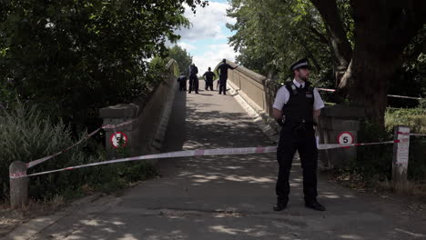 Un-Oficial-De-Policía-Hace-Guardia-En-Un-Cordón-Policial-Grabado-Mientras-Un-Equipo-Forense-Investiga-Un-Puente-Luego-De-Un-Incidente-De-Crimen-Con-Armas-Y-Cuchillos