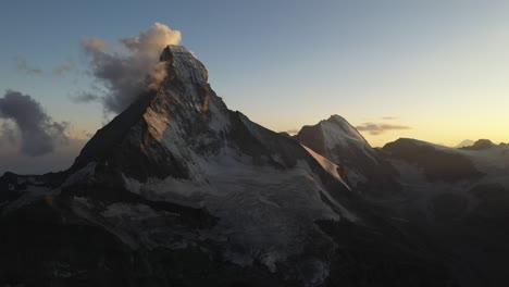 Drone-shot-moving-forward-with-moving-and-dancing-clouds-changing-form-and-shape-as-they-stick-and-stay-around-the-pointy-snowy-peak-of-the-mountain-Matterhorn-in-Switzerland-during-an-epic-sunrise