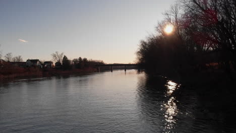 Bright-sun-shining-through-dark-trees-along-while-traveling-down-Susquehanna-river,-Aerial