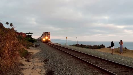 Statische-Ansicht-Des-Vorbeifahrenden-Amtrak-Zugs,-Während-Zwei-Mädchen-Bei-Bewölktem-Sonnenuntergang-Am-Rand-Der-Gleise-Fotografieren