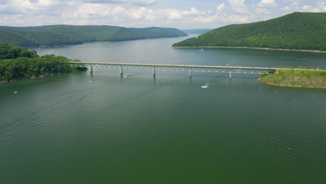 Aerial-drone-flying-forward-above-blue-and-green-lake-in-Pennsylvania-forest-in-the-summer-as-boats-and-jetskis-drive-over-water-and-cars-cross-the-bridge-over-the-lake