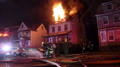 Firefighters-Arrive-On-Scene-and-Ready-Fire-Hoses-Next-to-a-Burning-Two-and-a-Half-Story-Home-at-Night