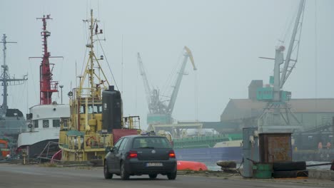 Hafenkräne-Beladen-An-Einem-Nebligen-Tag-Ein-Trockenfrachtschiff-Im-Hafen-Von-Liepaja,-Ein-Auto-Fährt-Vorbei,-Totale