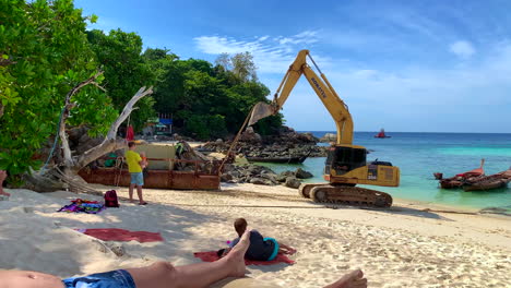 Eine-Aufnahme-Eines-Baggers,-Der-Einen-Benzin--Oder-Öltank-Am-Strand-Entlang-Schleppt,-Während-Männer-Daran-Arbeiten-Und-Touristen-Nebenher-Zuschauen