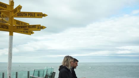 SLOWMO---Young-attractive-couple-looking-at-ocean-by-Stirling-Point-sign-in-Bluff,-New-Zealand