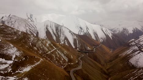 Brown-and-white-mountain-with-cold-frozen-ice-road-and-snow-covered-mountains-in-clouds-and-fog-in-background
