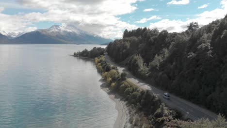 SLOWMO---Campervan-driving-along-the-coast-of-Lake-Wakatipu,-Queenstown,-New-Zealand-with-mountains-fresh-snow-in-background---Aerial-Drone