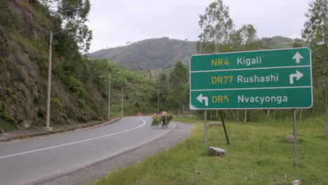 Schwenken-Sie-Nach-Rechts-Zum-Straßenschild,-Das-Nach-Kigali,-Ruanda-Führt,-Mit-Einer-Person-Auf-Einem-Fahrrad,-Die-Durch-Den-Rahmen-Fährt