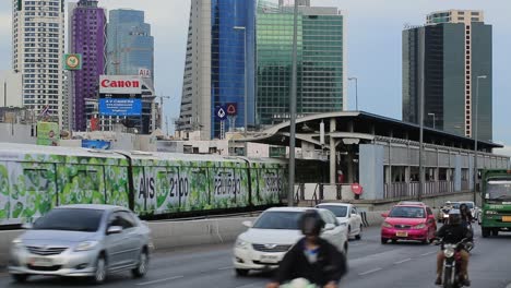 Bangkok,-Thailand---Transport-Vehicles-Moving-Fast-In-The-Highway-While-Train-Is-Coming-Out-Of-The-Station-With-High-Rise-Buildings-In-The-Background---Close-up-Shot