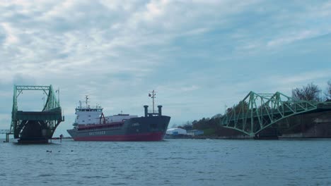 Blue-and-red-colored-cargo-ship-enters-the-port-via-an-open-Oskara-Kalpaka-swing-bridge-in-Liepaja-in-cloudy-autumn-day,-wide-shot