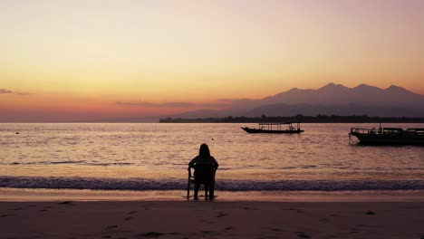 Silhouette-of-young-woman-sitting-on-chair,-watching-beautiful-sunset-reflecting-on-sea-surface-in-Bali