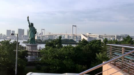 Réplica-De-La-Estatua-De-La-Libertad-De-Odaiba-En-La-Ciudad-De-Minato-Con-El-Puente-Colgante-Del-Arco-Iris-En-El-Fondo,-Toma-De-Mano