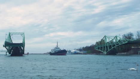 Un-Pequeño-Barco-Pesquero-Entra-En-El-Puerto-A-Través-De-Un-Puente-Oscilante-Oskara-Kalpaka-Que-Se-Cierra-En-Liepaja-En-Un-Día-Nublado-De-Otoño,-Plano-General