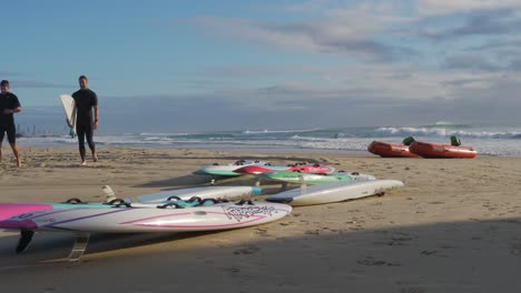 Tablas-De-Surf-Y-Botes-De-Rescate-Tirados-En-La-Arena-De-La-Playa---Dos-Surfistas-Activos-Caminando-En-La-Playa-Después-De-Surfear---Currumbin,-Costa-Dorada,-Australia