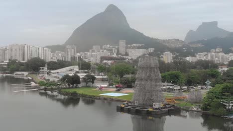 Panorámica-Aérea-Que-Muestra-El-árbol-De-Navidad-Flotante-Parcialmente-Construido-En-La-Orilla-Del-Lago-De-La-Ciudad-En-Río-De-Janeiro-En-Una-Mañana-Brumosa-Con-La-Montaña-De-Los-Dos-Hermanos-En-El-Fondo