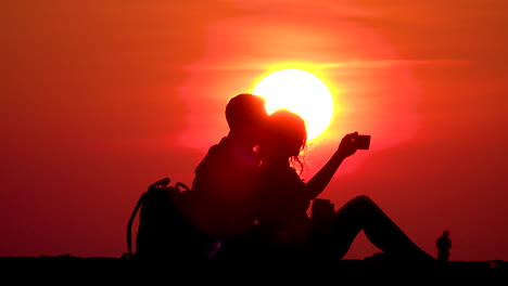 A-couple-sitting-at-North-Avenue-Beach-in-Chicago-taking-selfies-on-a-cellphone-in-front-of-a-bright-yellow-sun-with-orange-sky