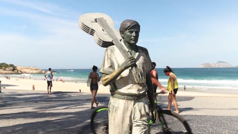People-passing-by-bronze-statue-of-composer-Tom-Jobim-holding-a-guitar-over-his-shoulder-on-Ipanema-boulevard-with-palm-tree-and-Arpoador-beach-and-rock-in-the-background